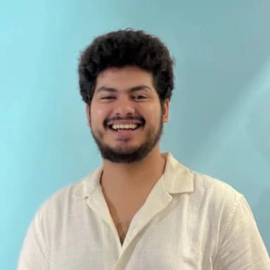 Portrait of Geetansh Saini, smiling, in a cream shirt against a light blue wall.